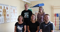 Fünf Therapeuten posieren in einem Therapieraum mit anatomischen Postern.