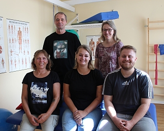Fünf Therapeuten posieren in einem Therapieraum mit anatomischen Postern.